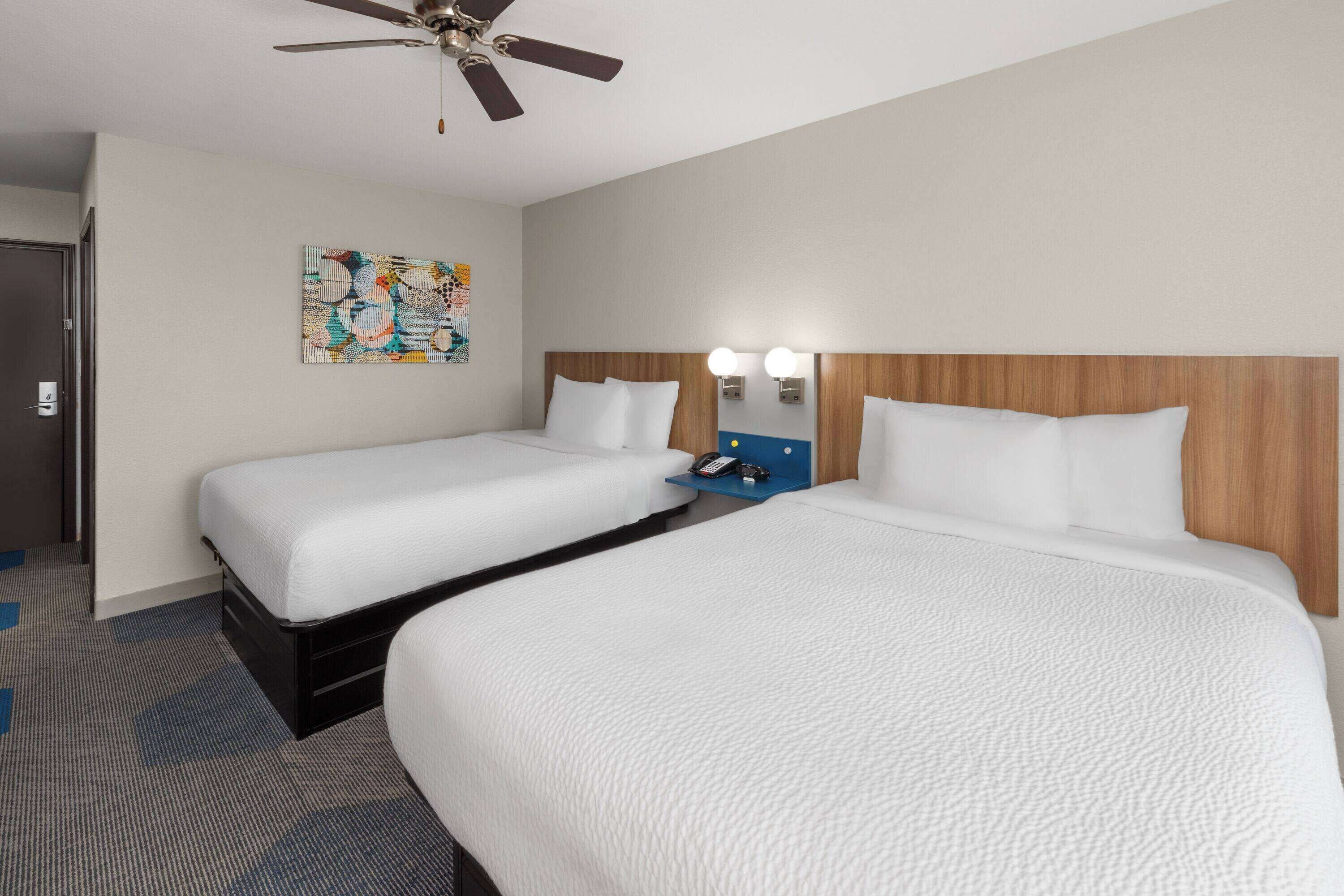 A hotel room with two beds, a ceiling fan, a wall-mounted light, and a door.