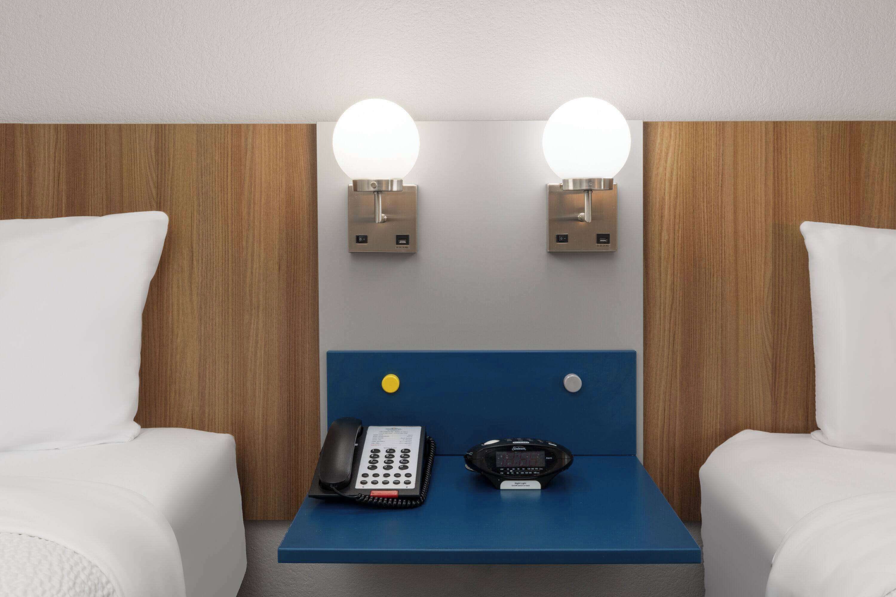 A hotel room with a wooden headboard, two bedside lamps, a desk with a telephone and a digital device, and two white pillows.