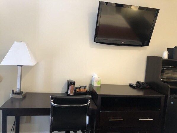 A hotel room with a flat-screen TV mounted on the wall, a desk with a lamp, a chair, a coffee maker, and a microwave. - Seaside Inn (San Francisco)