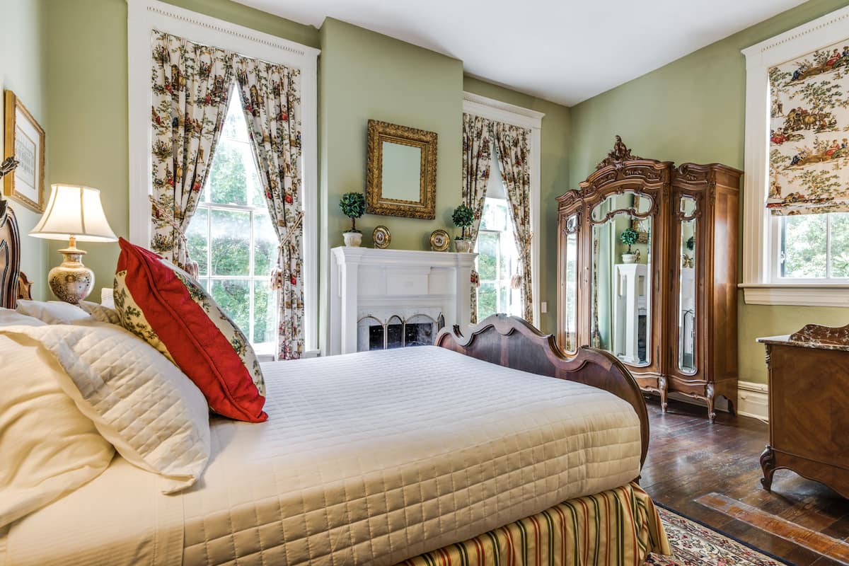 Quarto com cama grande, lareira, espelho e janela com cortinas florais.