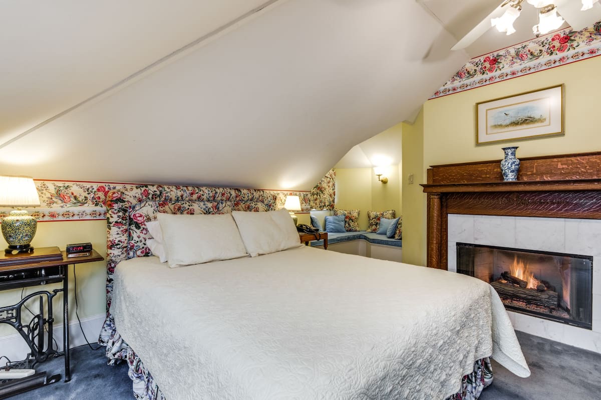 Quarto com uma cama grande, lareira, escrivaninha com abajur e uma cadeira.