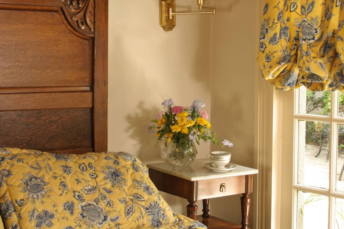 Quarto com cabeceira de madeira, mesa de cabeceira com abajur, um vaso com flores e uma janela com cortinas.