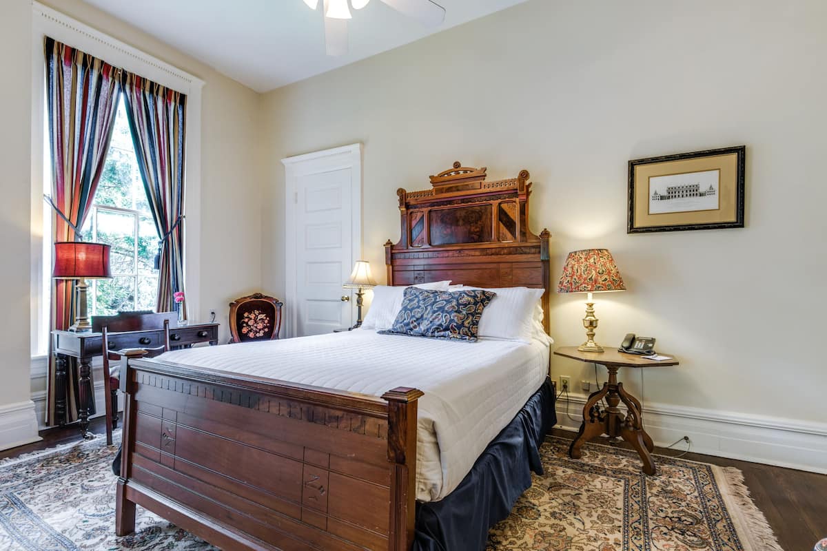Quarto com cama de madeira, escrivaninha, cadeira, abajur e quadro pendurado na parede.