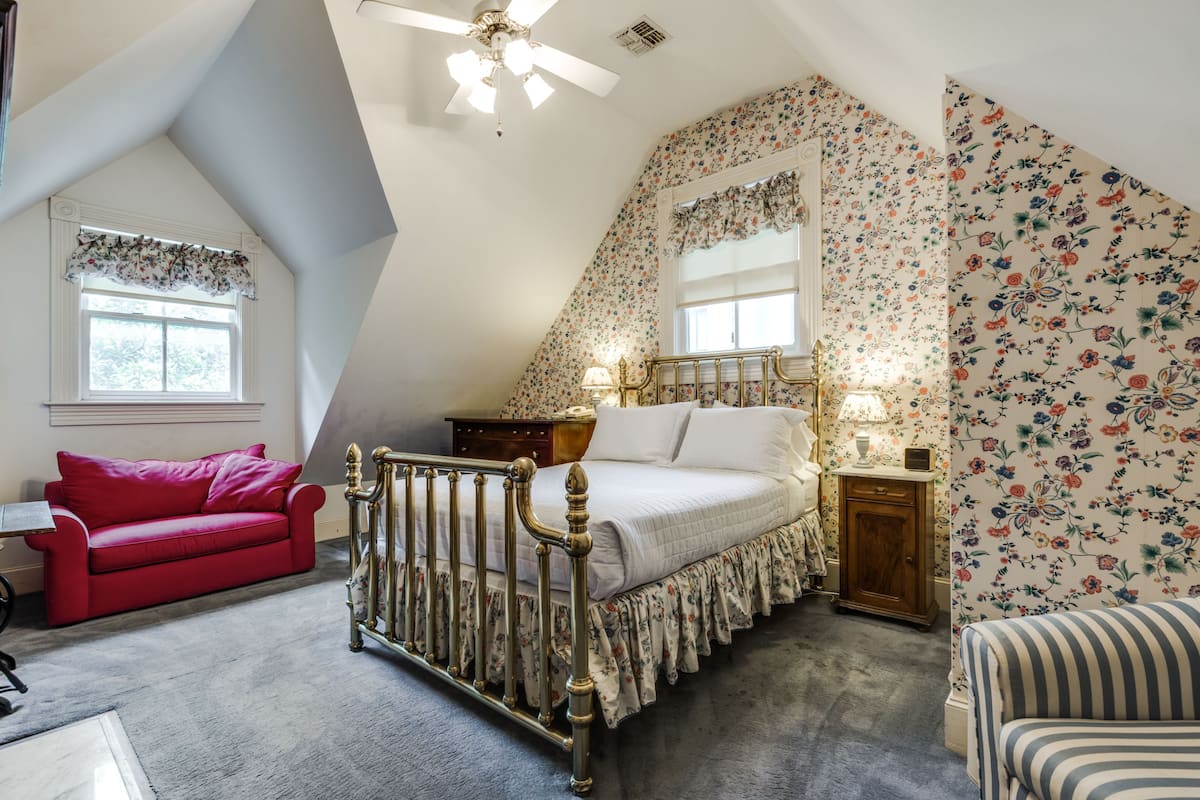 Um quarto com teto inclinado, uma cama com colcha florida, um sofá vermelho e uma mesa de cabeceira com abajur.