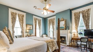 A bedroom with a large bed, a ceiling fan, a fireplace, a desk with a chair, and a television. - Noble Inns (San Antonio)