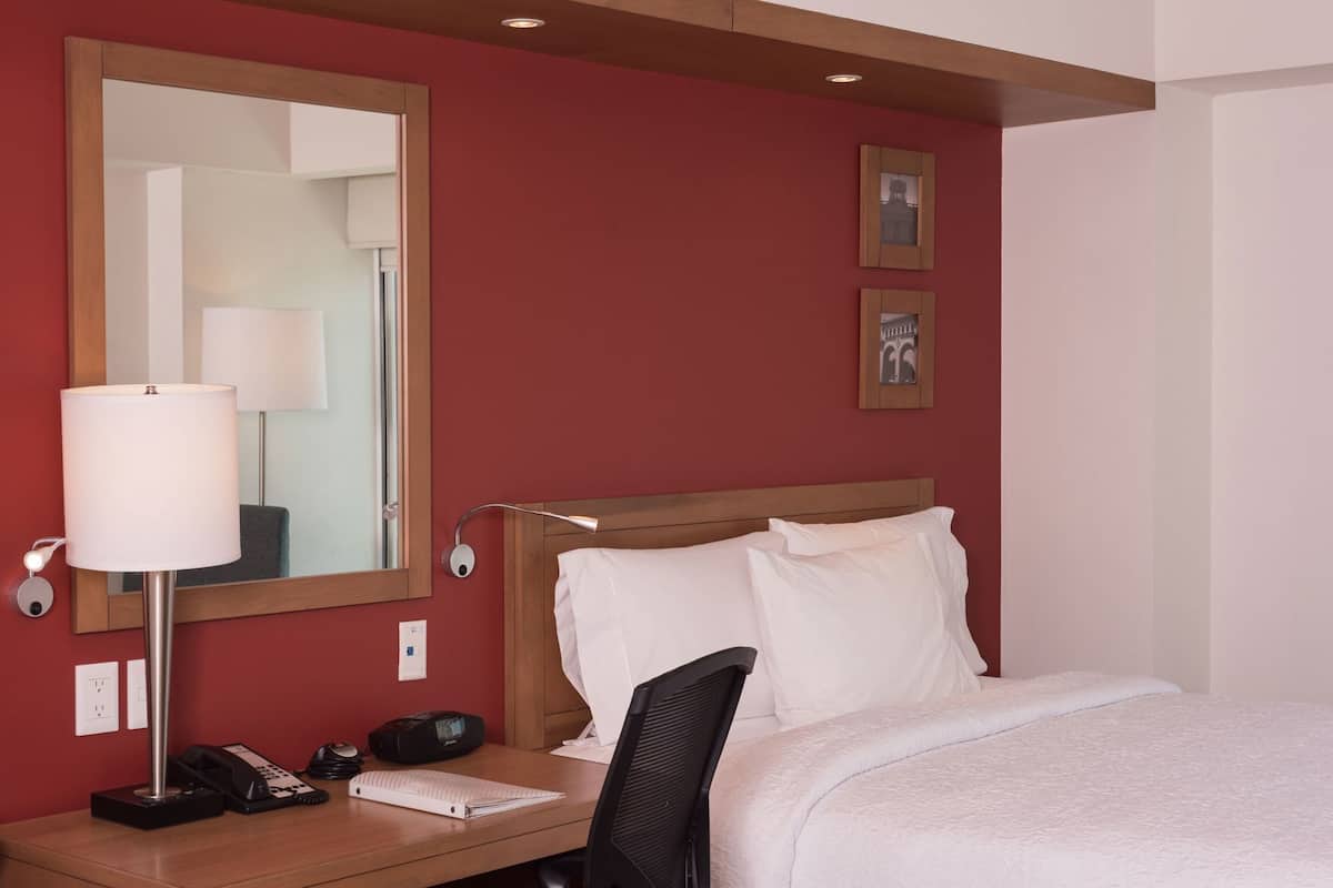 Habitación de hotel con cama, escritorio con lámpara, teléfono, espejo y cuadros en la pared.