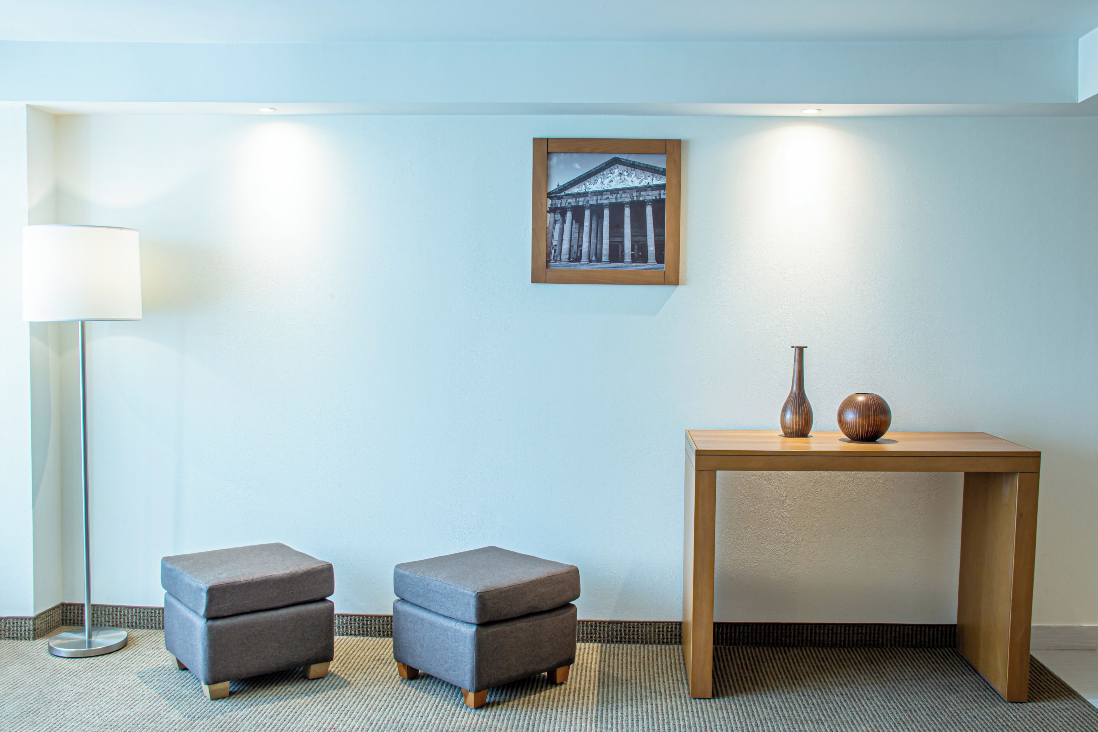 A room with a light blue wall, a framed picture of a building, a wooden table with two vases, and two grey ottomans.