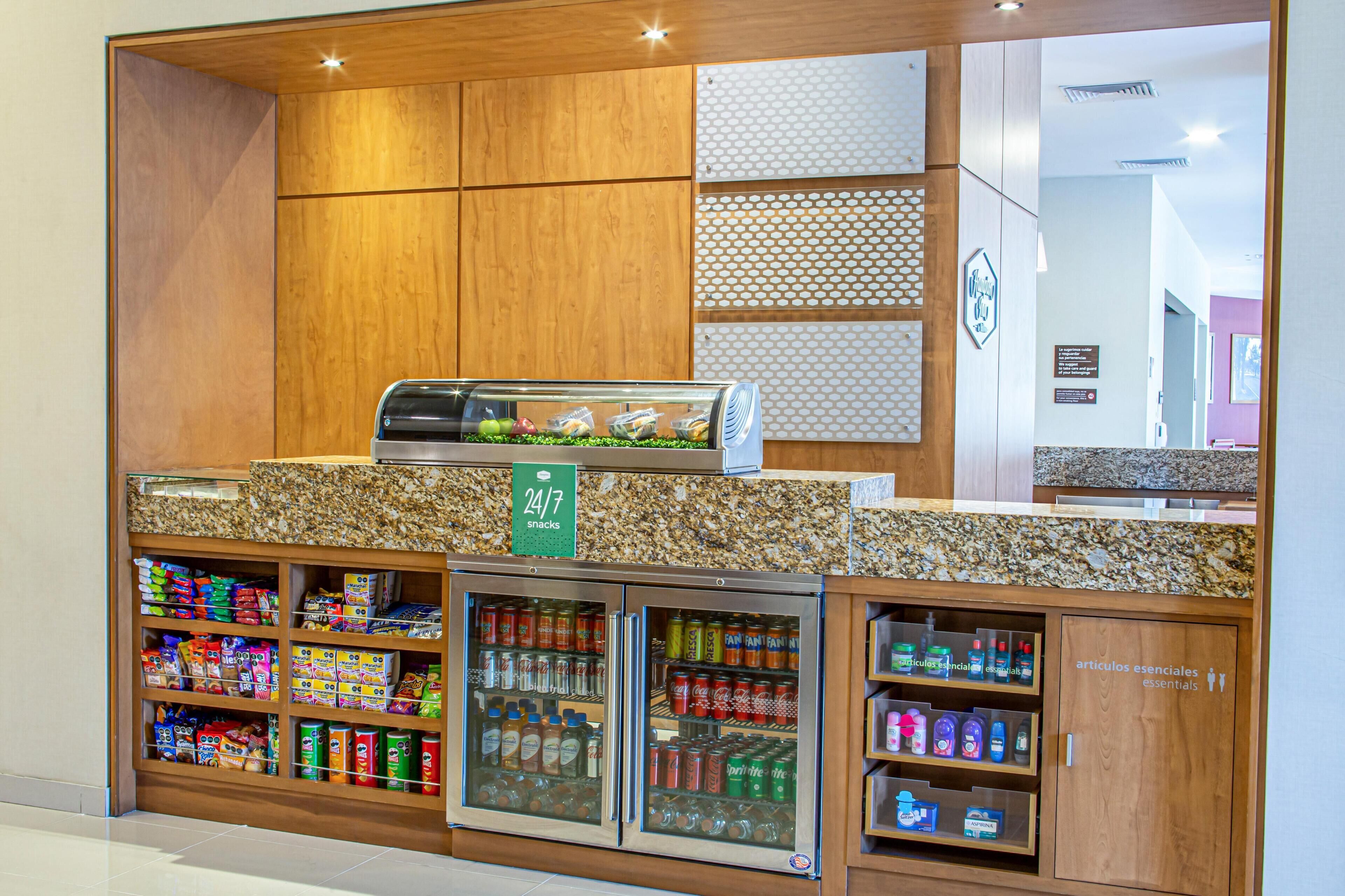 Uma área de lanches com autosserviço, com diversas opções de alimentos e bebidas embalados, uma máquina de café e um aviso indicando disponibilidade 24 horas por dia.