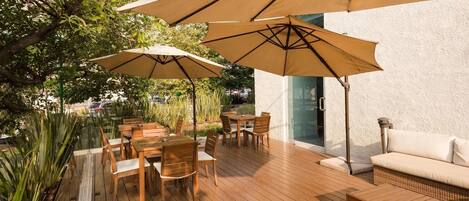 Un patio al aire libre con mesas y sillas de madera, sombrillas color beige y un sofá blanco.