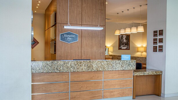 Un mostrador de recepción de hotel con diseño de paneles de madera, un cartel que dice "Hampton Inn by Hilton" y un letrero de "Hilton Honors".