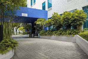 A building entrance with a blue overhang, paved walkway, and lush greenery. - Hampton Inn by Hilton Guadalajara/Expo (Guadalajara)
