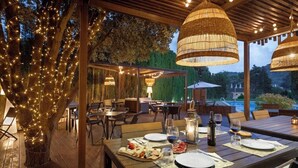 Un área de comedor al aire libre con mesas dispuestas para una comida, luces colgantes y una estructura de pérgola.