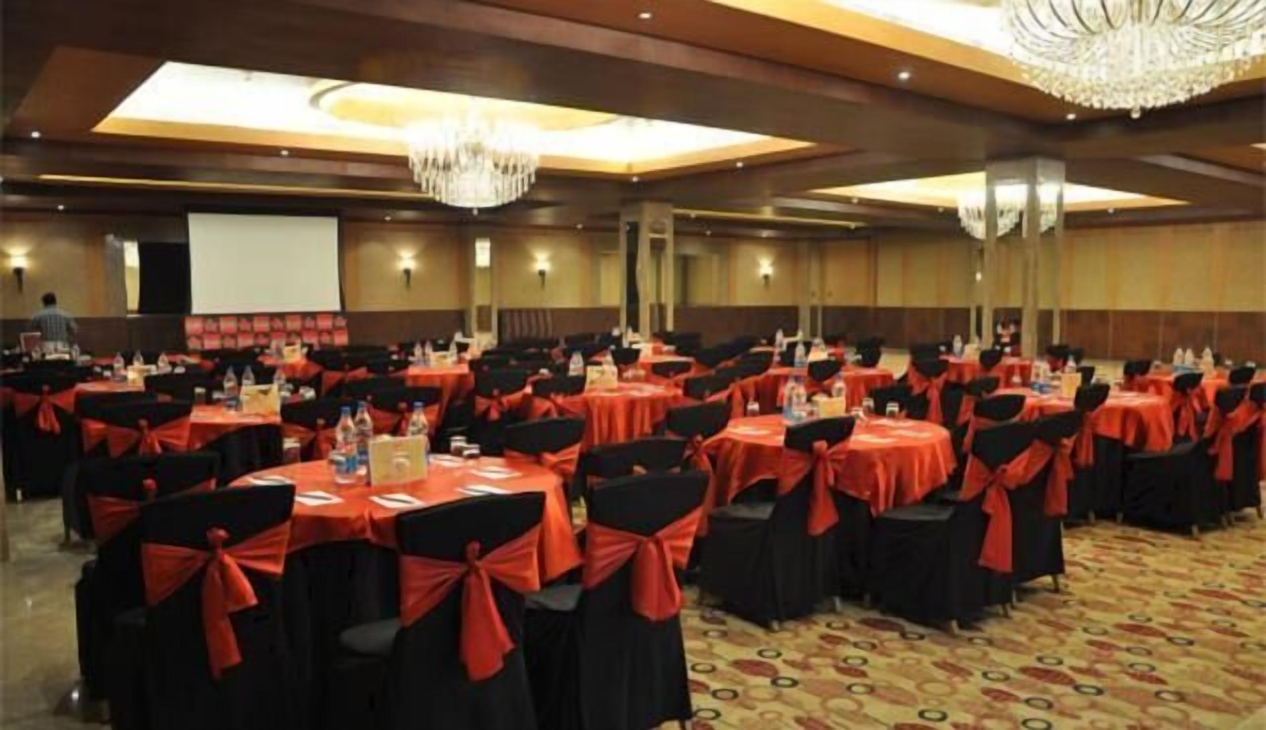 A banquet hall with round tables, black chairs with red sashes, and a chandelier.