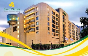 A multi-story hotel building with a modern architectural design, featuring balconies and a prominent glass tower. - Savoy Suites Manesar (Gurugram)