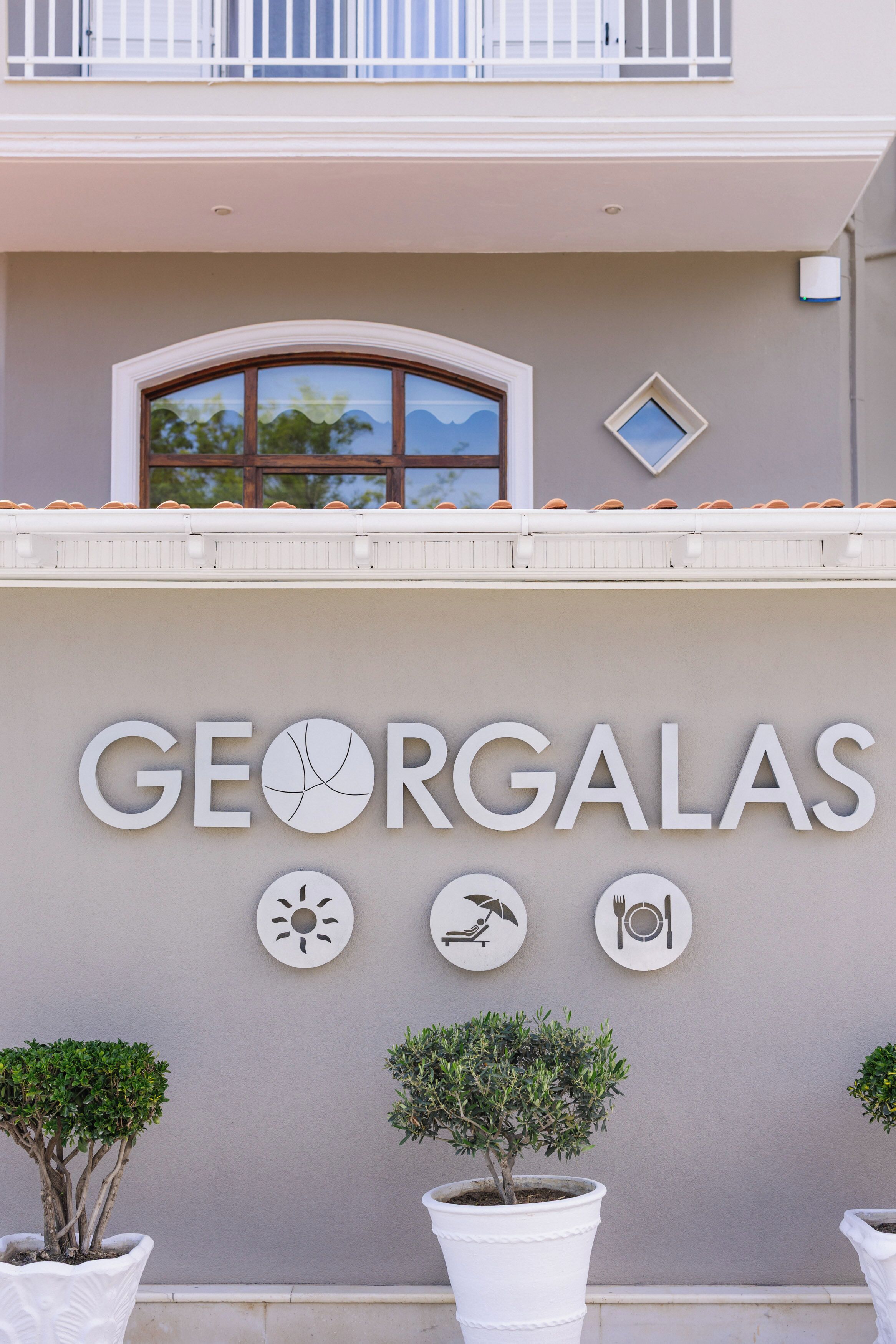 Un edificio con el nombre 'GEORGALAS' destacado en la fachada, acompañado de símbolos que representan sol, descanso y gastronomía.