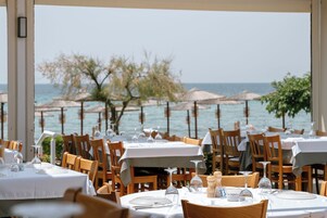 Restaurante con mesas dispuestas para comer, con vistas a un cuerpo de agua con sombrillas y árboles.