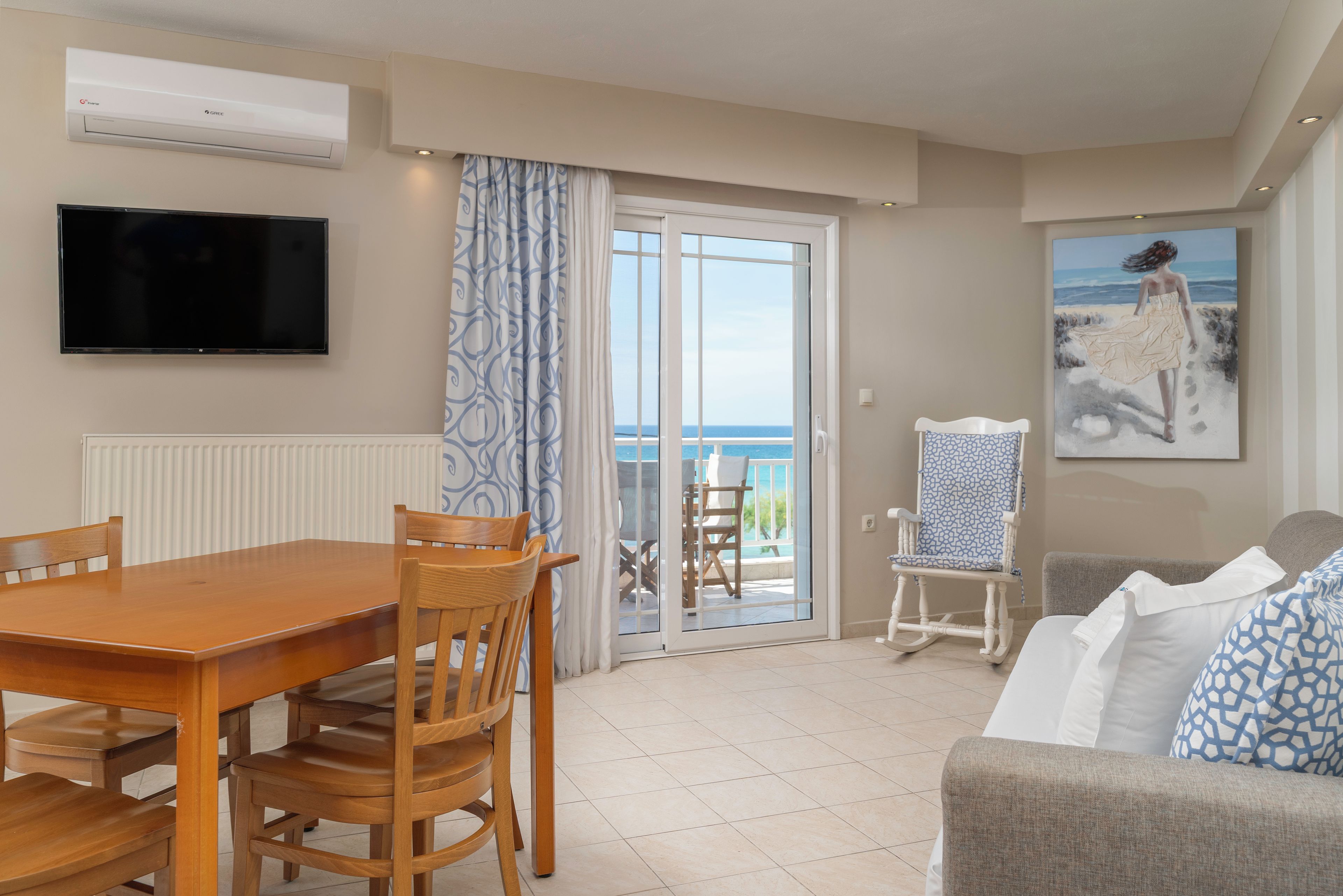 Una sala de estar con una mesa y sillas de madera, una televisión, un cuadro y vista al mar a través de un ventanal.