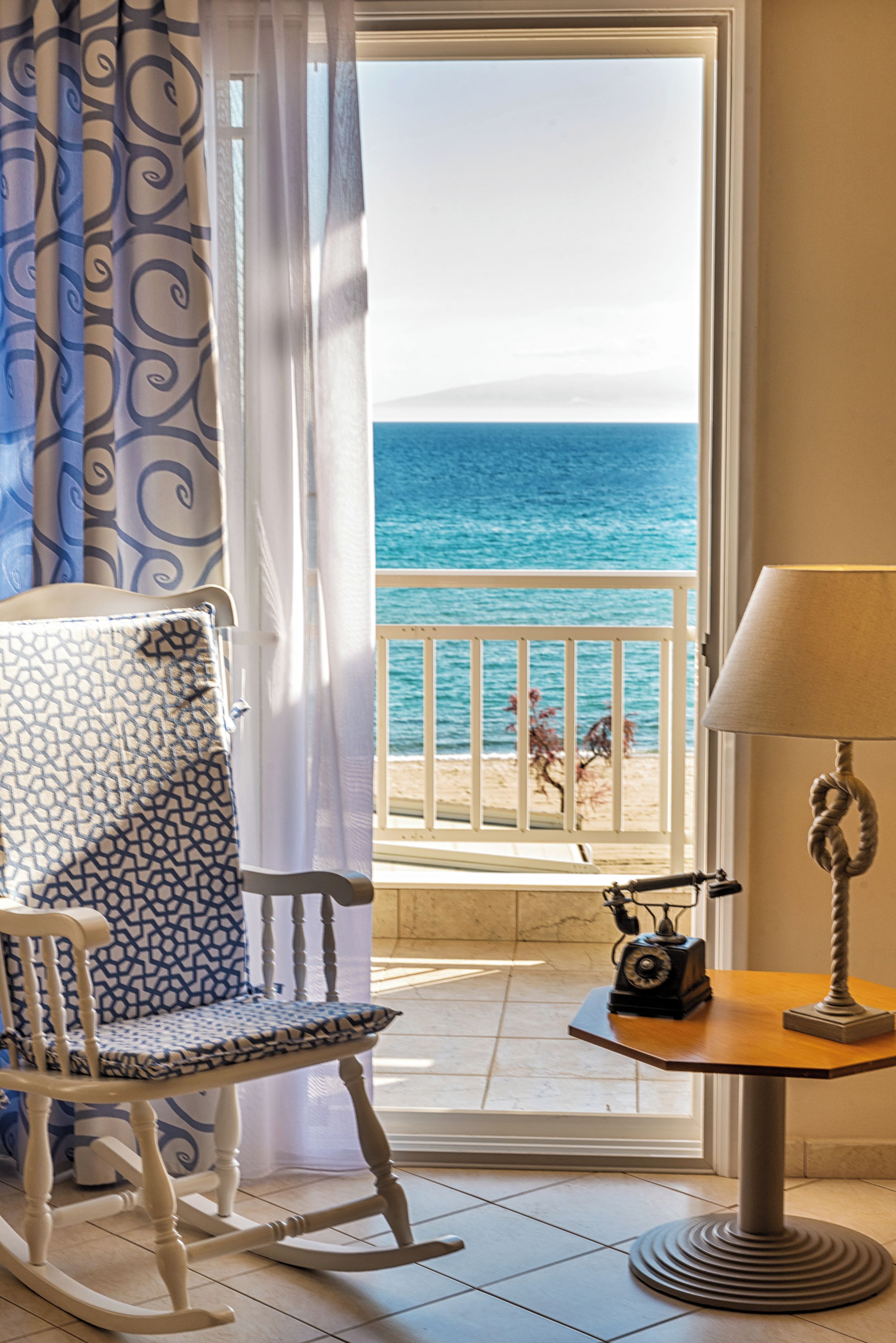 A room with a rocking chair, a table with a lamp, and a view of the ocean through a window.