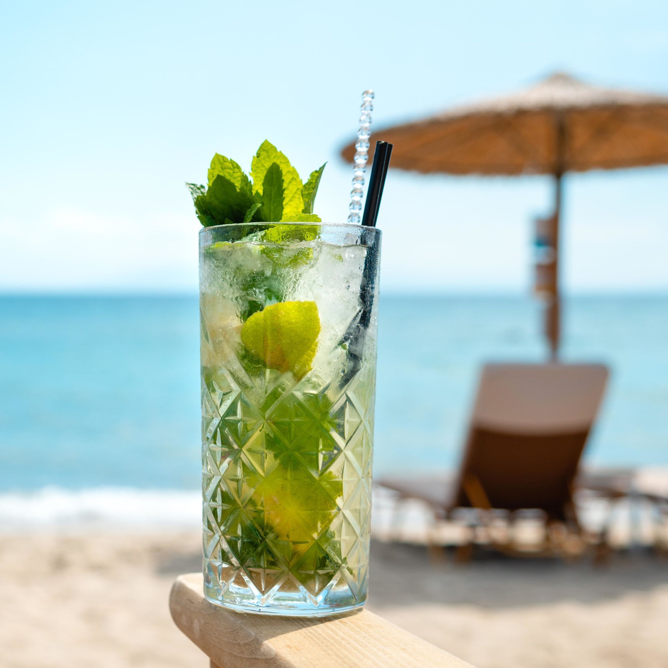 Un vaso transparente con hielo, lima y hojas de menta, con una pajita, sobre una superficie de madera, al lado de una playa con una silla y una sombrilla al fondo.