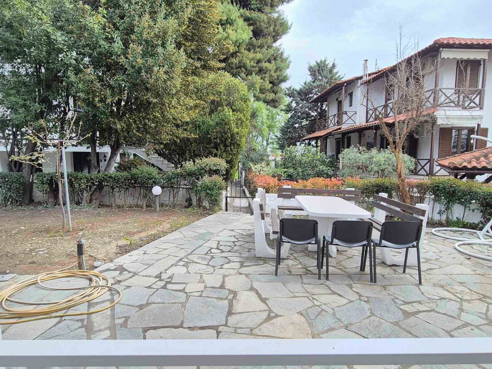 An outdoor patio with stone flooring, a table with chairs, and a garden with trees and shrubs.