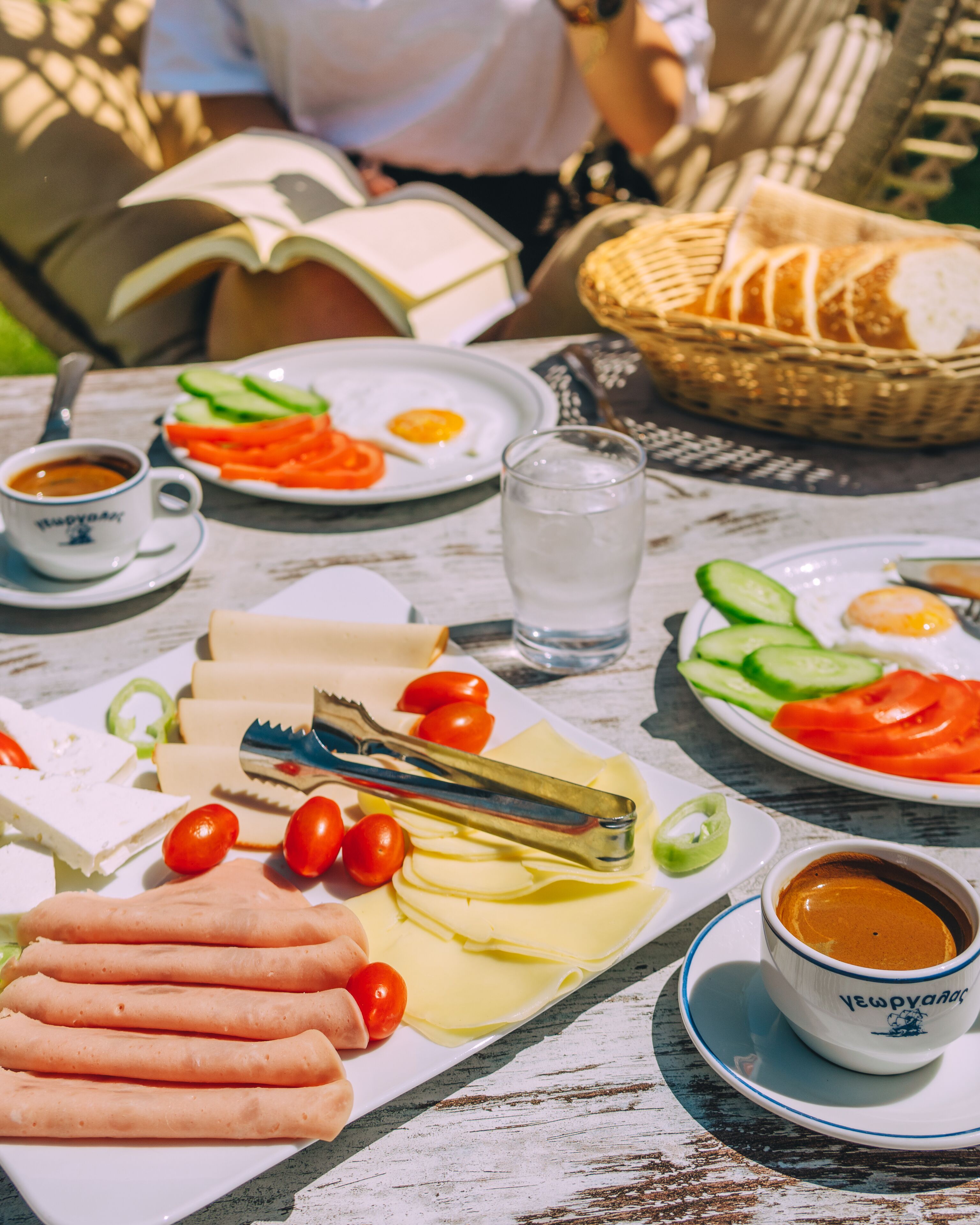 Una mesa servida con diversos alimentos como queso, tomates, pepinos y pan, junto con tazas de café y un vaso de agua.
