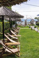 Filas de sillones de madera bajo un techo de paja, una estructura de piedra y vista al mar.