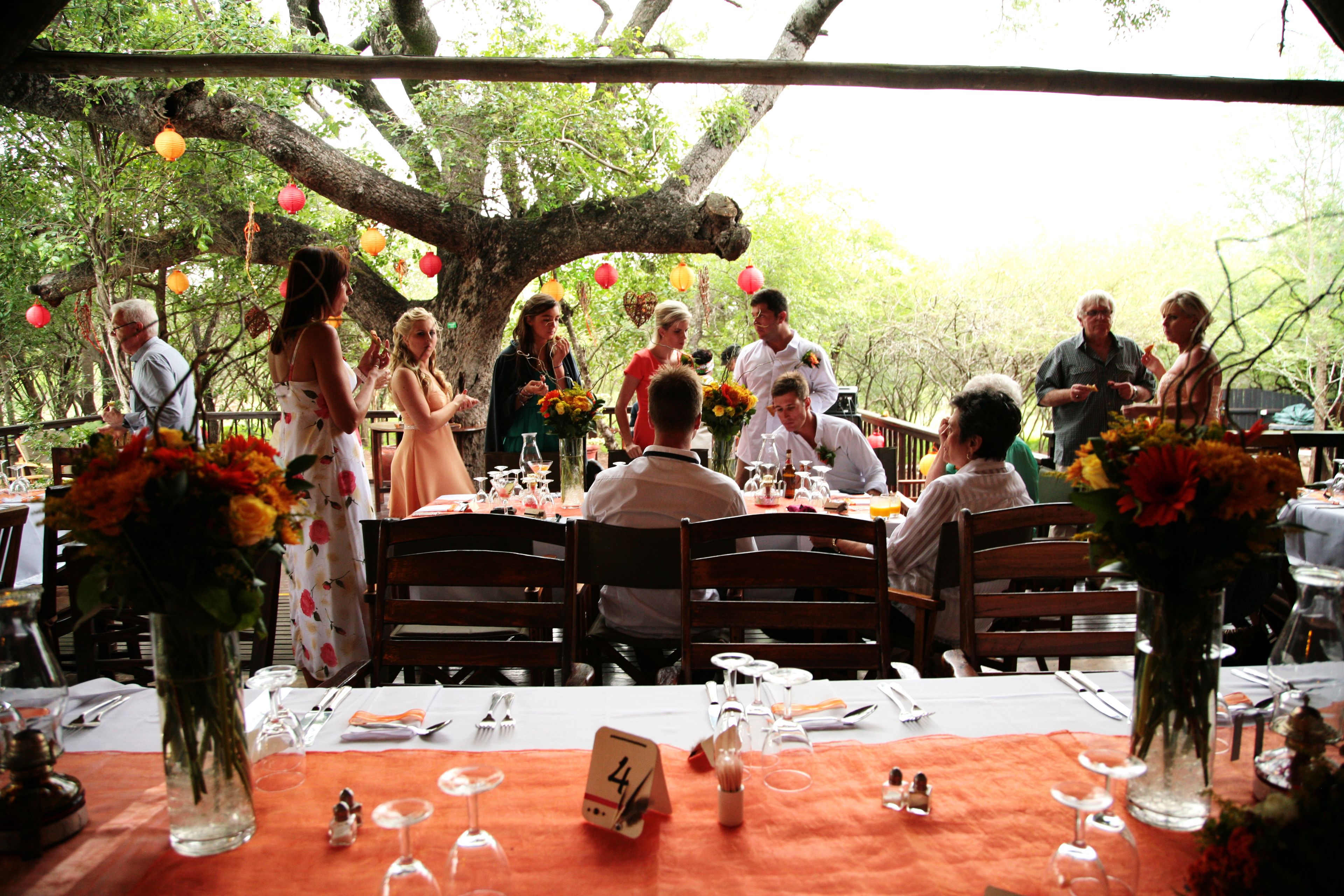 Un espacio de comedor al aire libre con mesas dispuestas para una comida, rodeado de árboles y decorado con faroles colgantes.