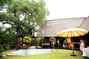 Relax by the pool surrounded by lush greenery and trees, with tables and chairs for dining or socializing.