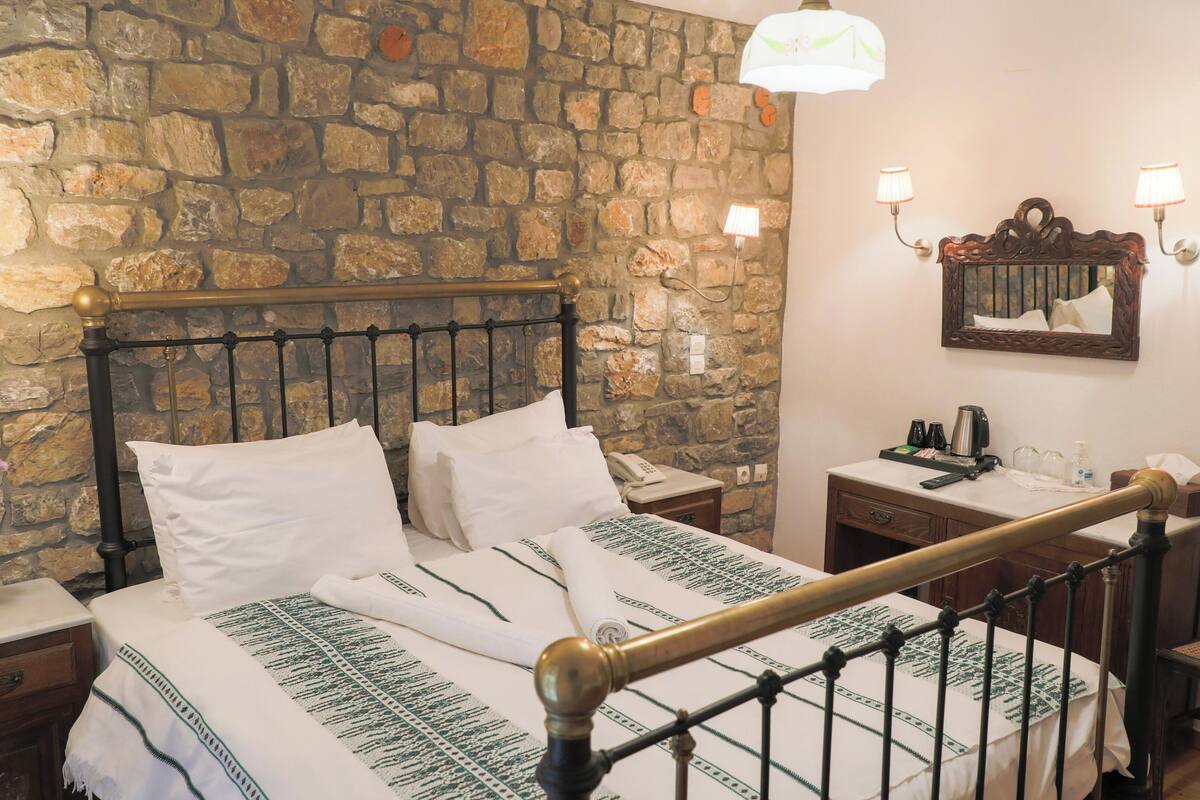 Ein Zimmer mit Steinwand, einem Bett mit weißen Leinen, einem Nachttisch mit Lampe, einem Holzstuhl und einem Spiegel an der Wand.