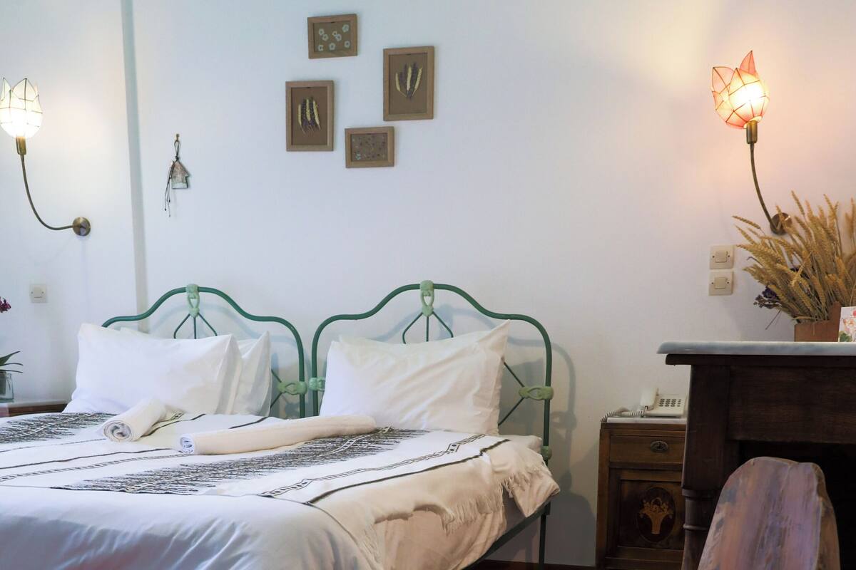 Ein Schlafzimmer mit einem grünem Metallbettgestell, weißer Bettwäsche, einem hölzernen Stuhl, einem Nachttisch mit Lampe und an der Wand befestigter Kunst.