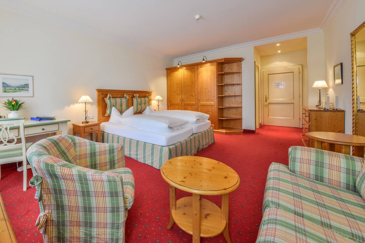 Habitación de hotel con cama, dos sillones a cuadros, una mesita redonda, un armario de madera y un escritorio con lámpara.