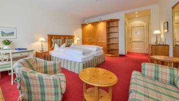 Une chambre d’hôtel avec un lit, deux fauteuils à motifs écossais, une petite table ronde, une armoire en bois et un bureau avec une lampe.