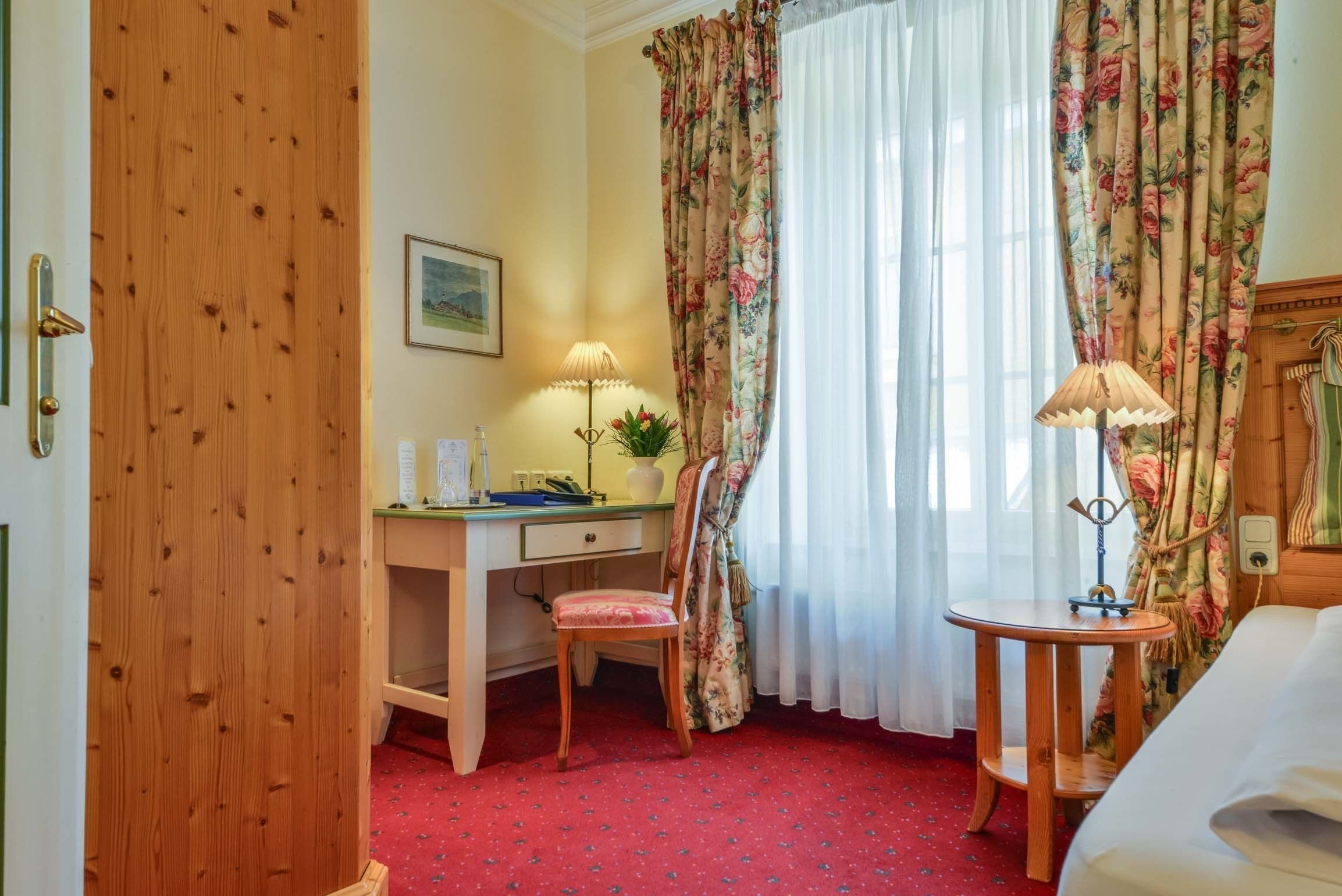 Ein Hotelzimmer mit Holztür, Schreibtisch mit Lampe, Stuhl, rundem Tisch und Bett.