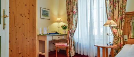 A hotel room with a wooden door, a desk with a lamp, a chair, a small round table, and a bed.