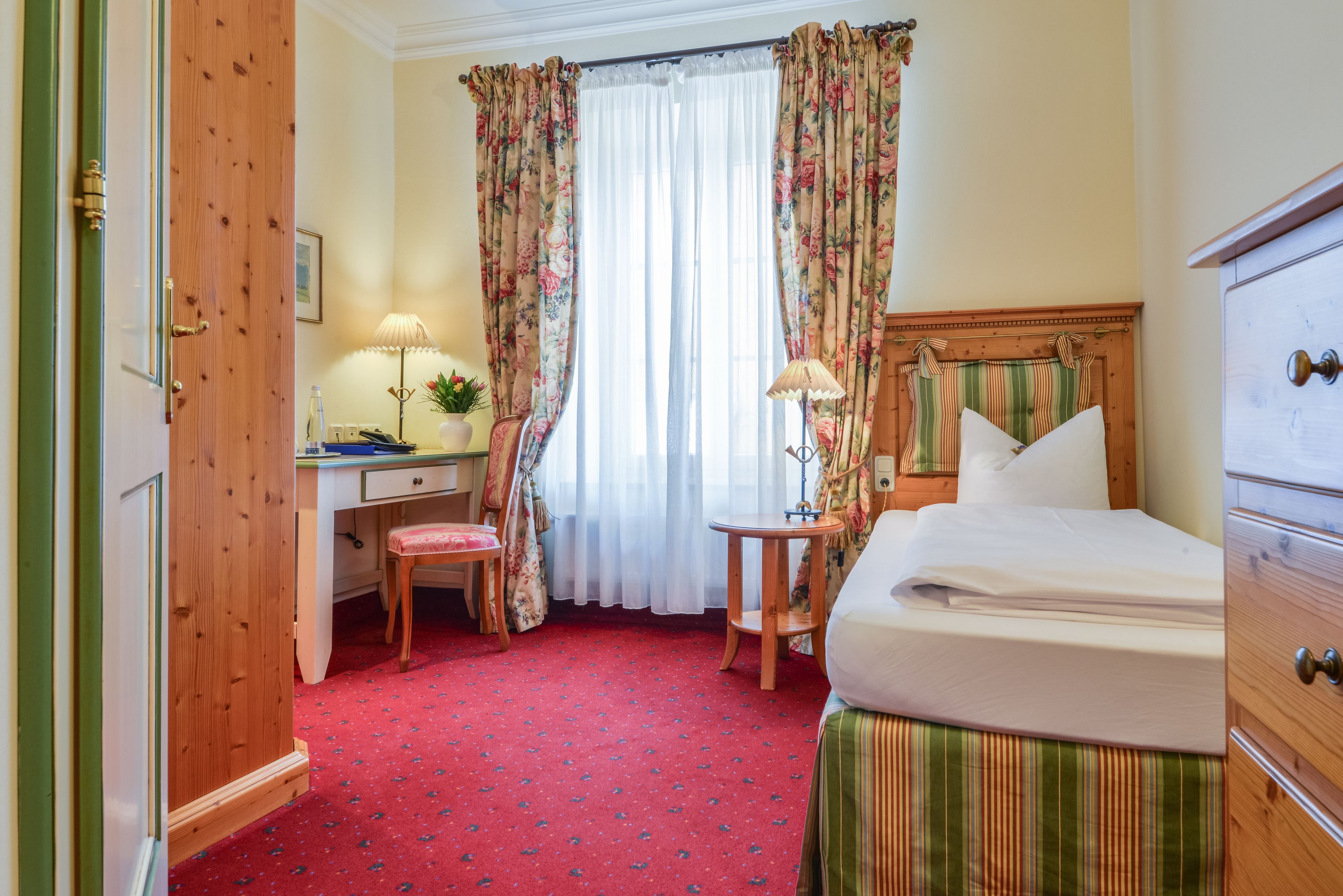 A hotel room with a wooden door, a desk with a lamp, a chair, a bed with a striped bedspread, and a window with floral curtains.