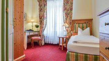 A hotel room with a wooden door, a desk with a lamp, a chair, a bed with a striped bedspread, and a window with floral curtains.