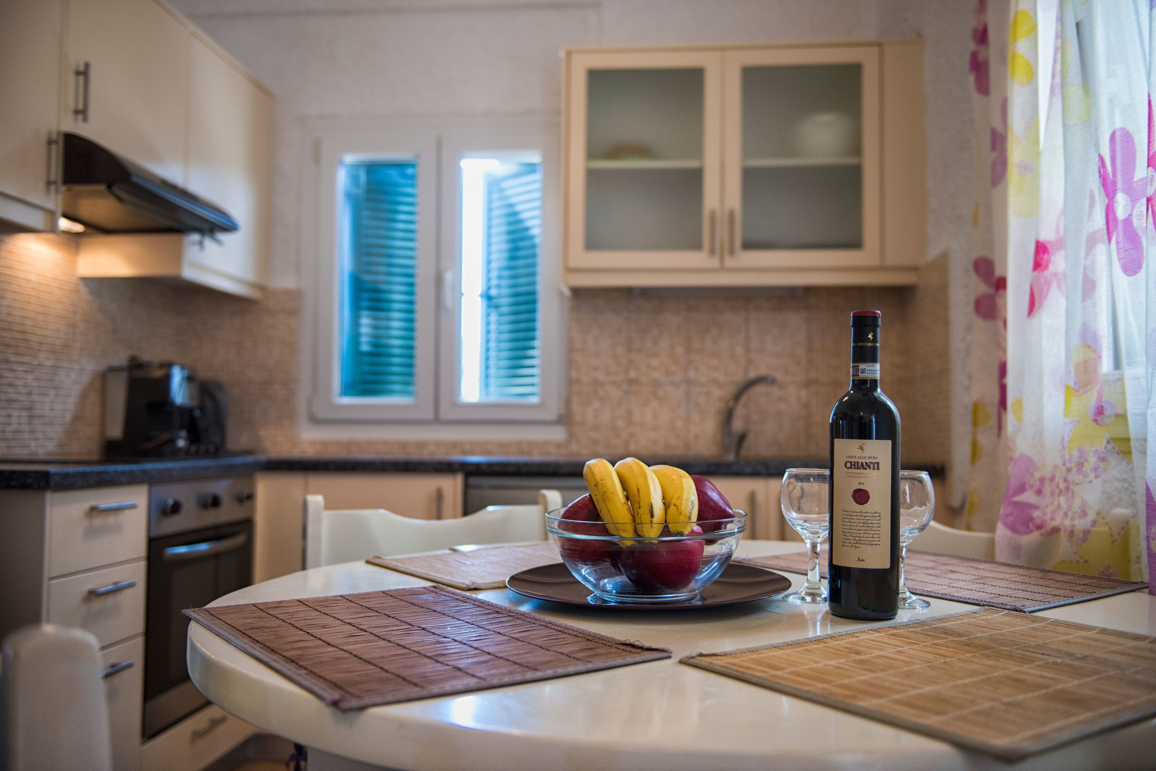 A kitchen with a dining table, a bowl of fruit, a wine bottle, and a window with blinds.