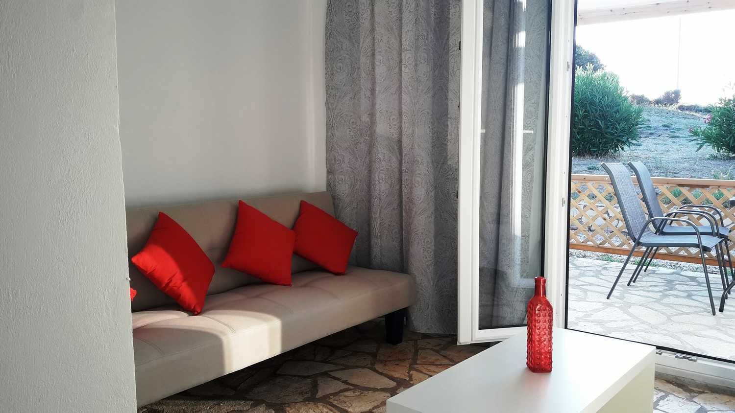Een woonkamer met een beige bank, rode kussens, een glazen deur naar buiten en een rode fles op een witte tafel.