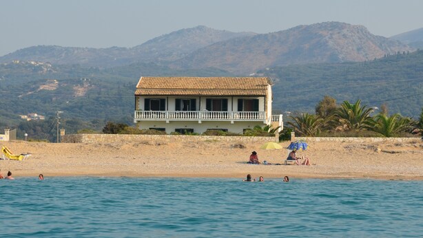 Una propiedad frente a la playa con un edificio grande, una playa de arena y montañas al fondo.