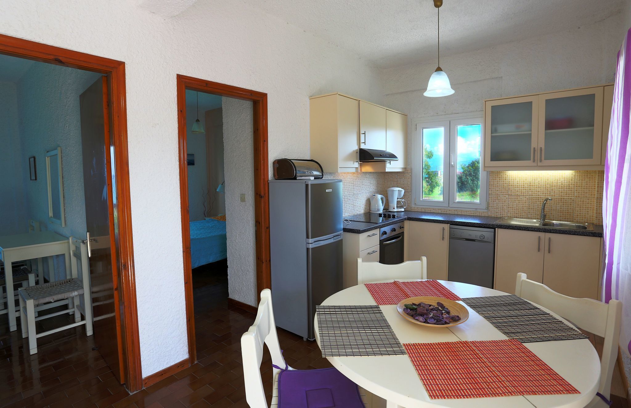 A compact kitchen with a refrigerator, stove, and sink. There is a dining table with placemats and a bowl of fruit.