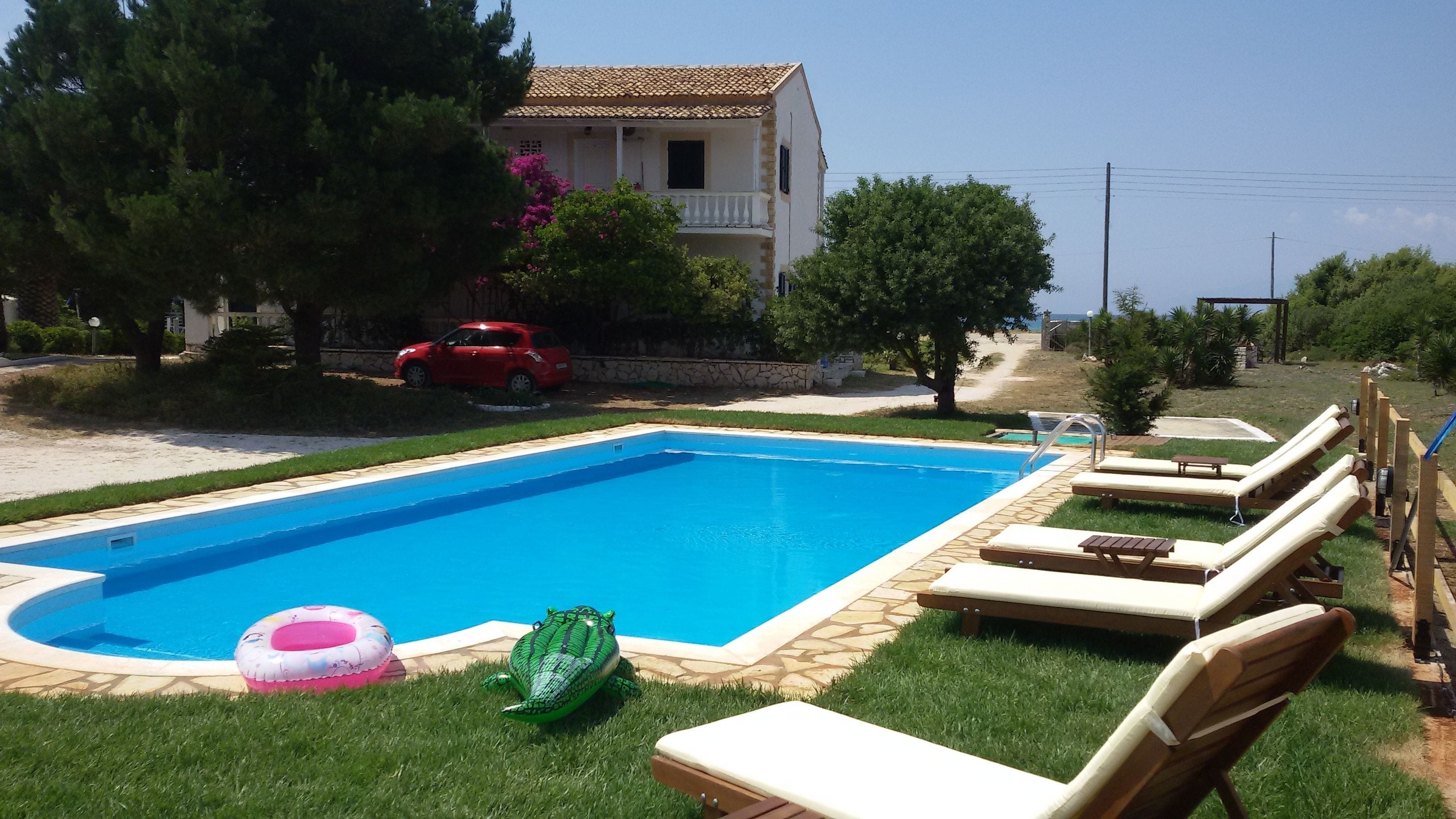 A swimming pool with lounge chairs, a red car, and a house in the background.
