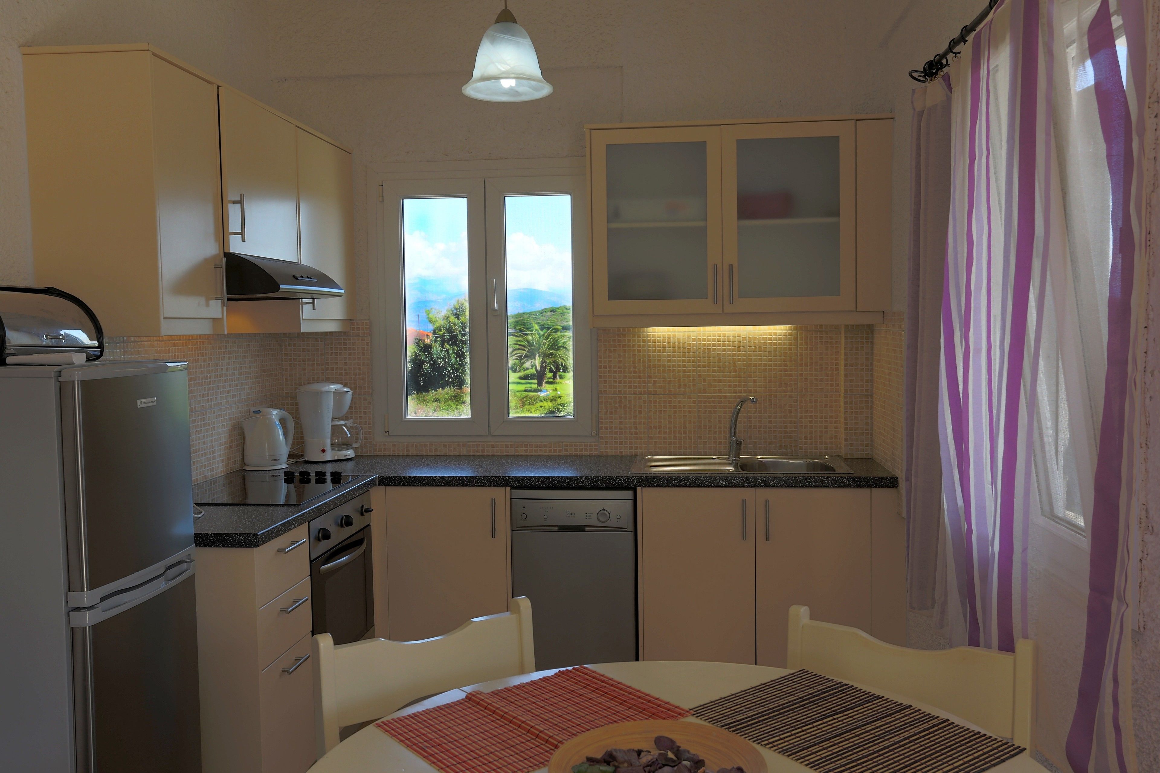 A compact kitchen with a refrigerator, stove, and dishwasher, a dining table with chairs, and a window with a view of greenery.