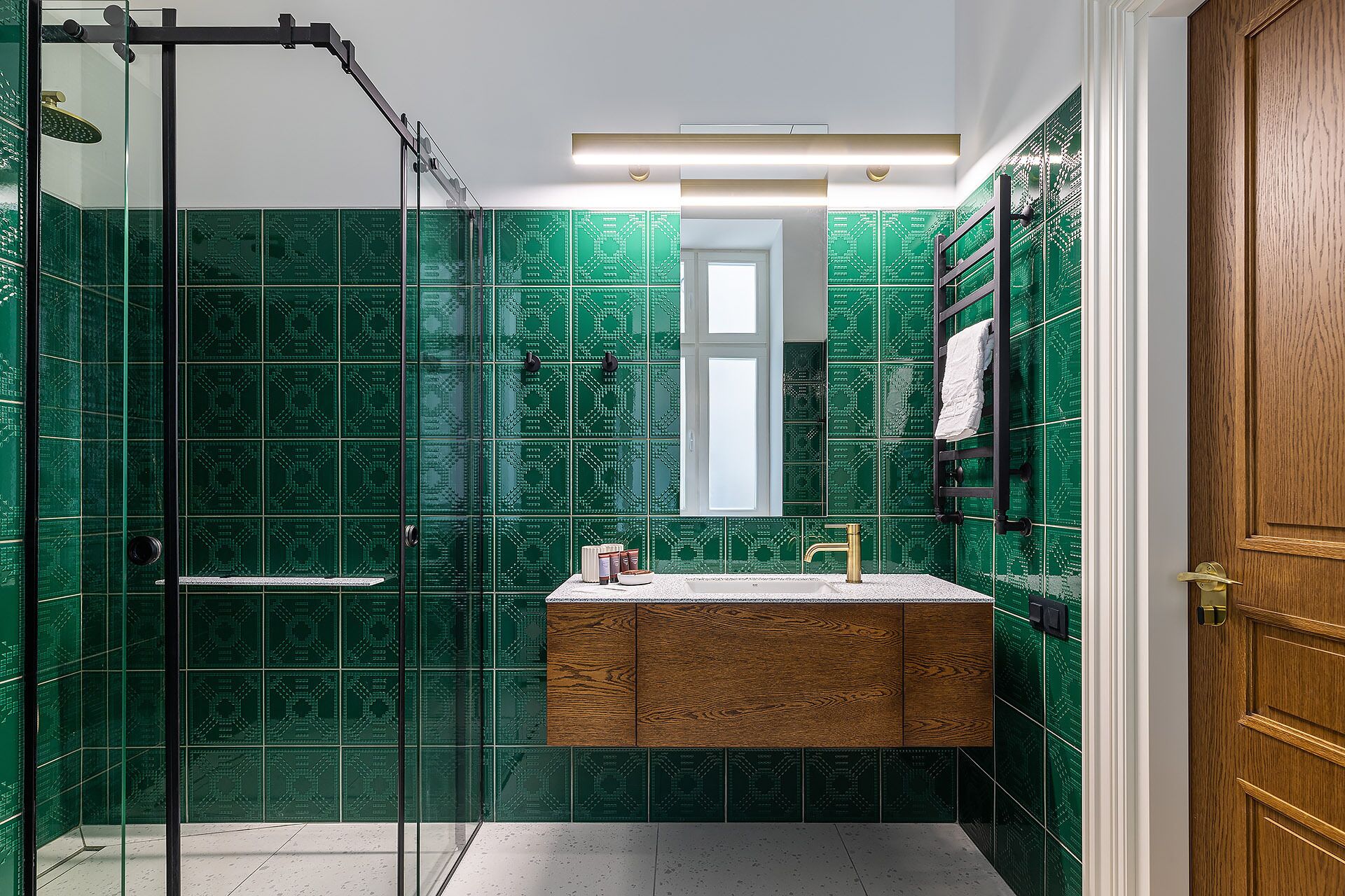 Ein Badezimmer mit grün gemusterten Fliesen, einem hölzernen Waschtisch, einer Glasdusche und einer Holztür.