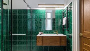 Ein Badezimmer mit grün gemusterten Fliesen, einem hölzernen Waschtisch, einer Glasdusche und einer Holztür.