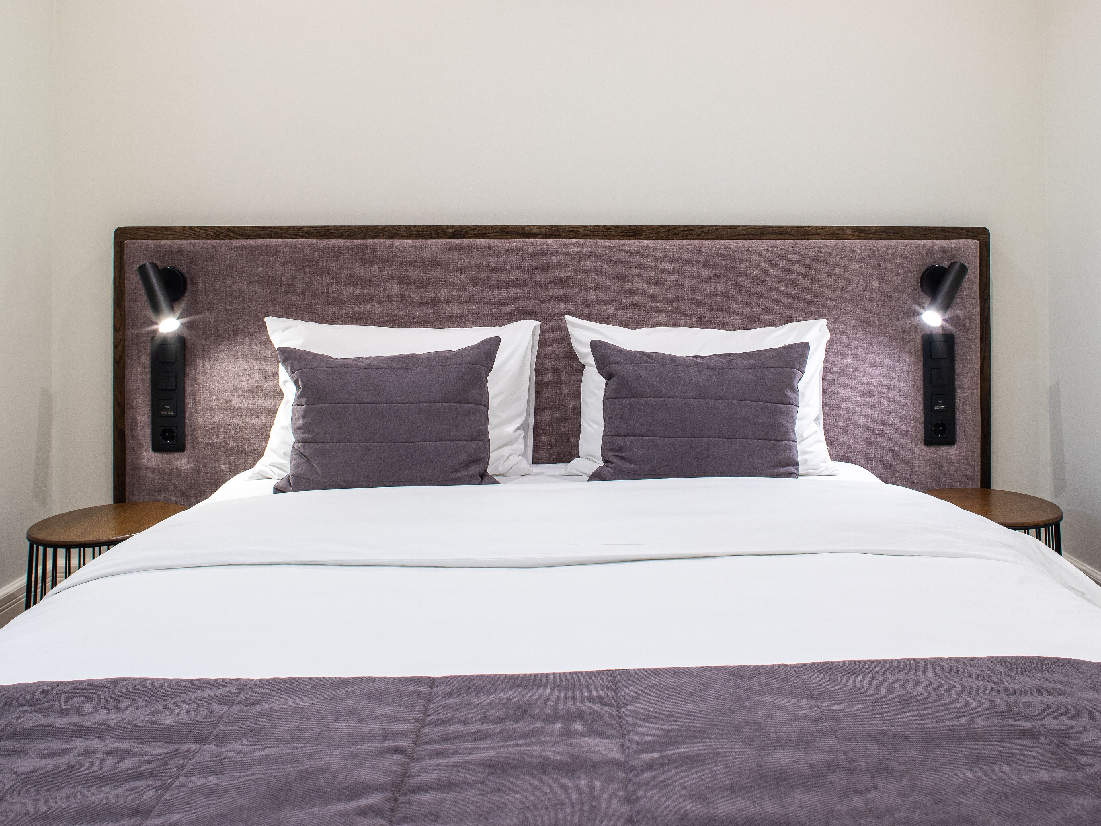 A neatly made bed with a white duvet and two dark pillows, flanked by two bedside tables with lamps.
