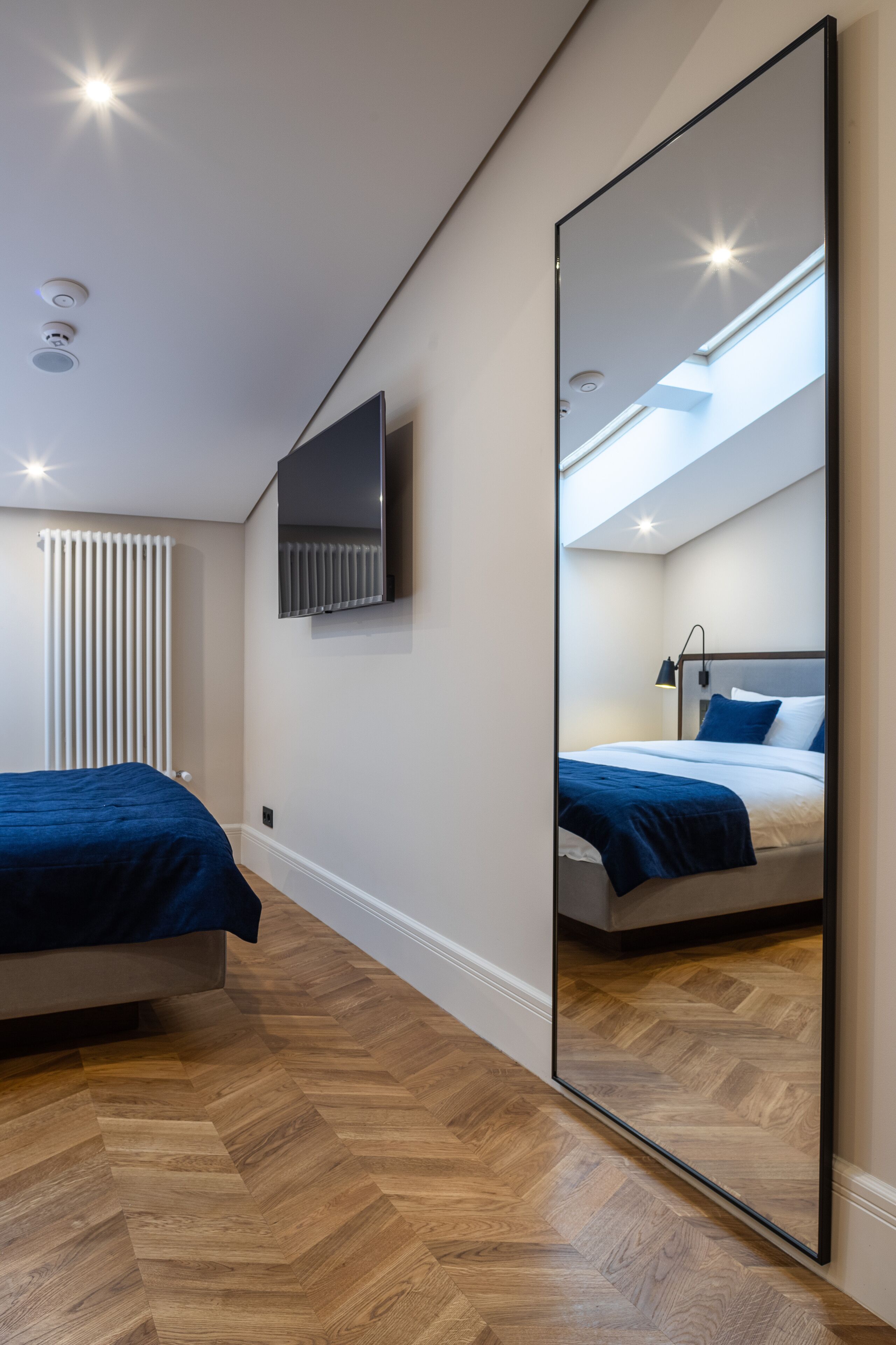 A modern bedroom with a large mirror, a bed with blue bedding, a TV mounted on the wall, and a wooden herringbone floor.