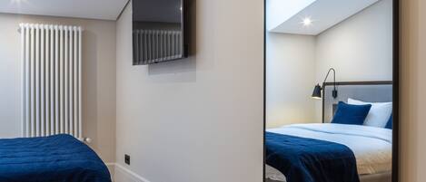 A modern bedroom with a large mirror, a bed with blue bedding, a TV mounted on the wall, and a wooden herringbone floor.