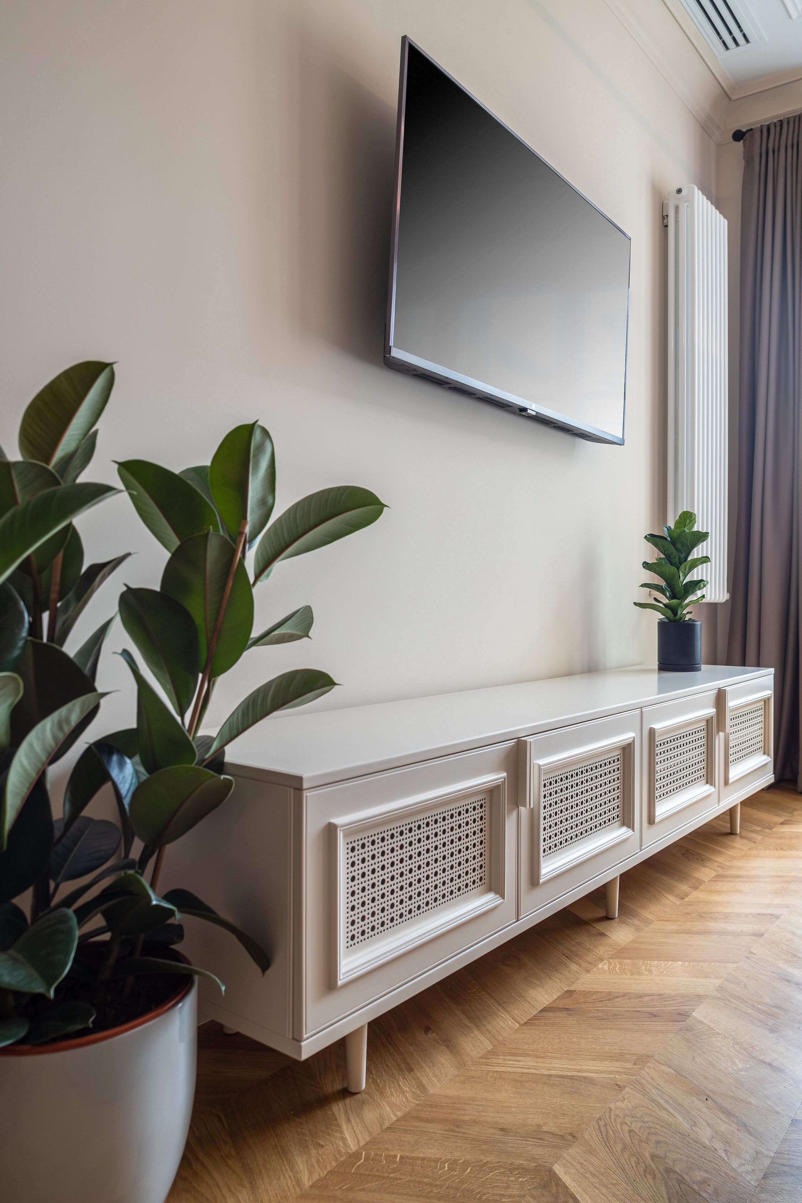 A modern living room with a flat-screen TV mounted on the wall, a white console table with decorative panels, and a potted plant.