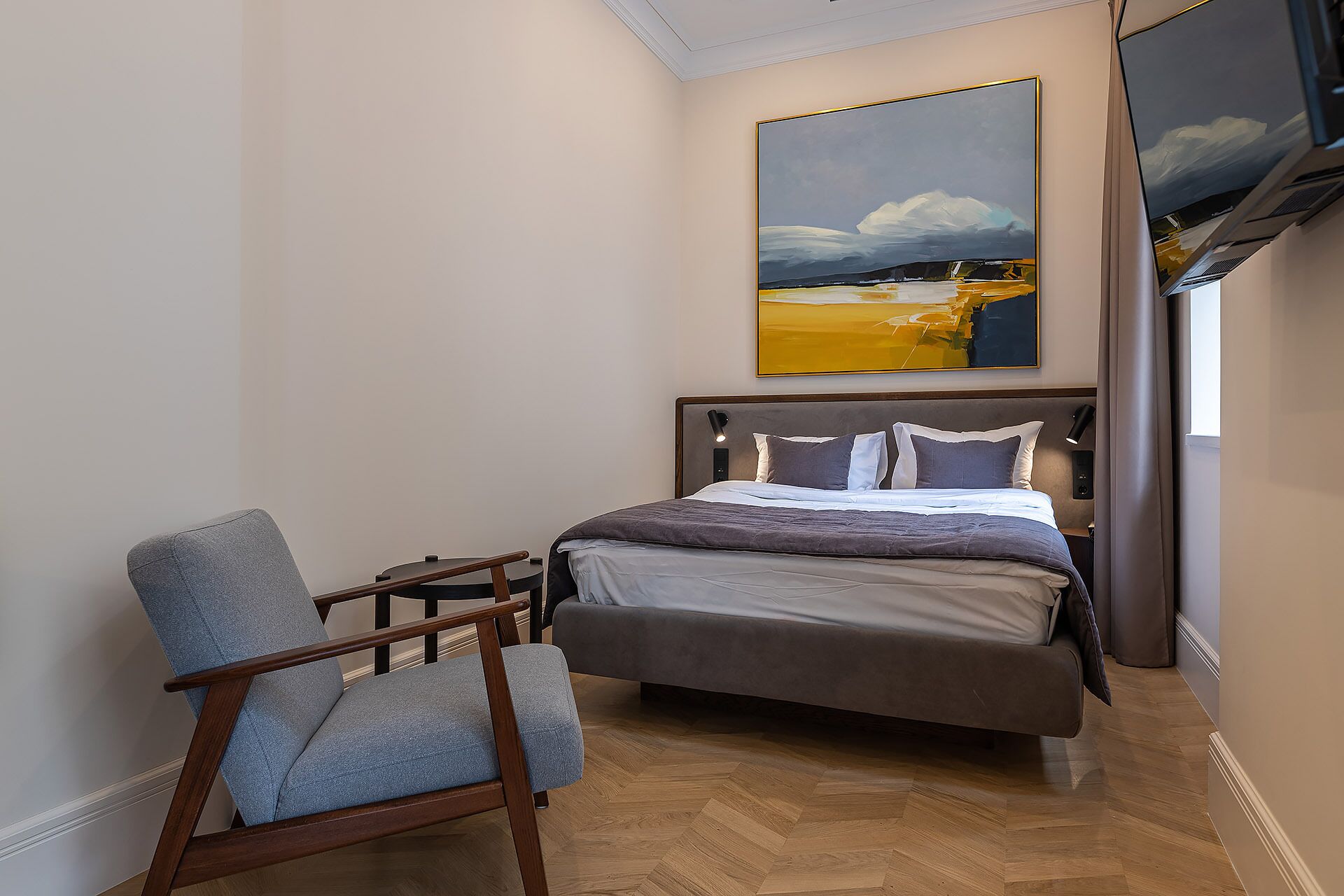 Un dormitorio con cama, una silla, un cuadro y un televisor en la pared.