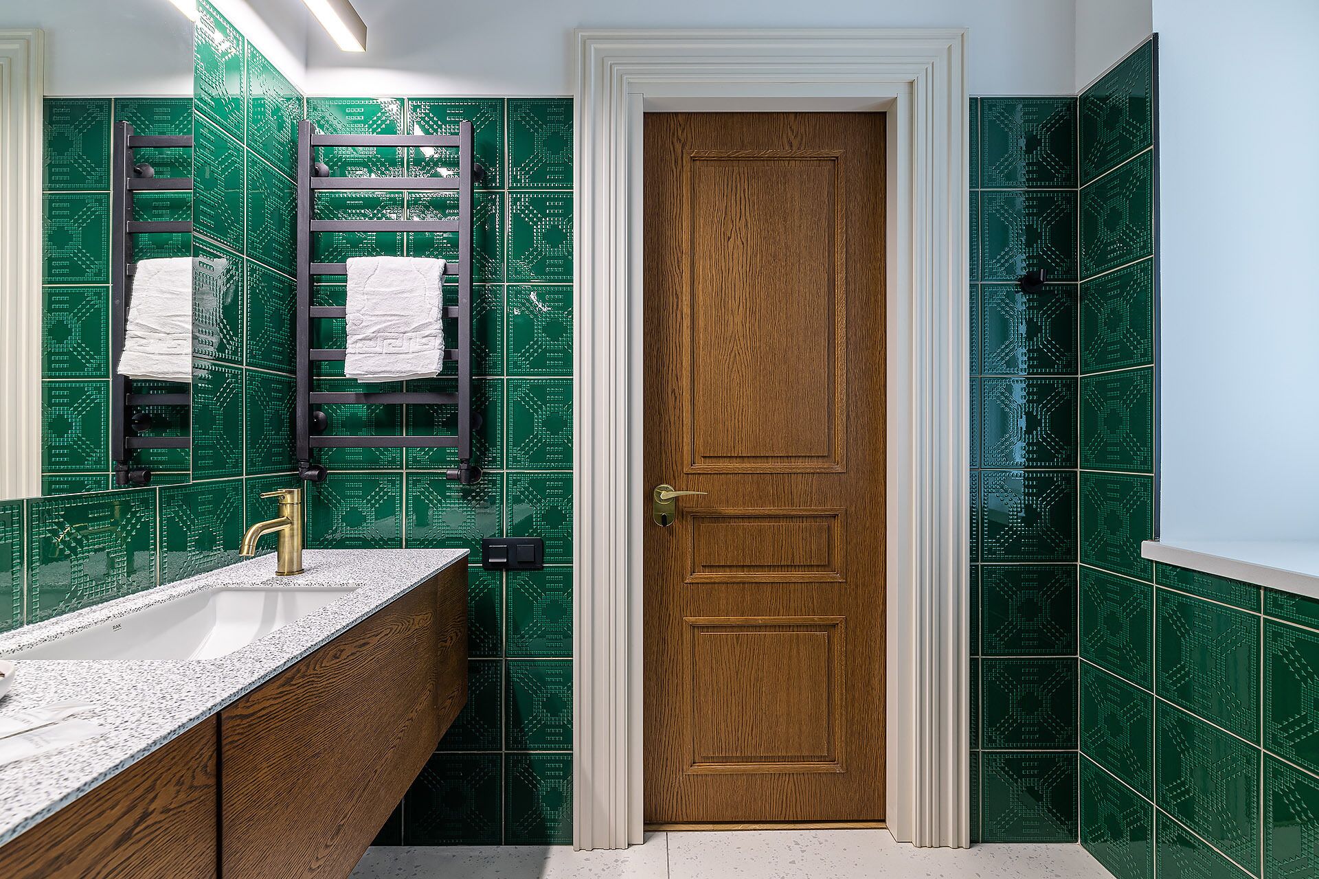 A bathroom with a wooden door, green patterned tiles, a towel rack, and a sink with a granite countertop.
